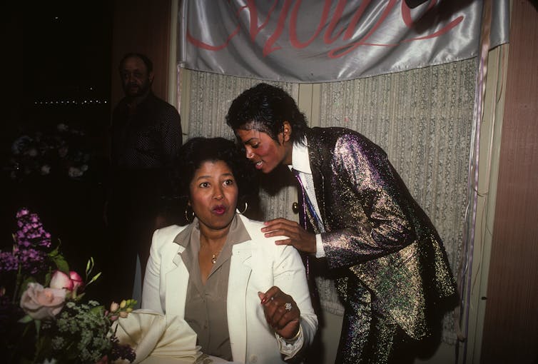 Katherine Jackson, wearing a white jacket over an olive green shirt, sits at a table covered with flowers while her son Michael Jackson, dressed in a sparkly suit, stands behind her.